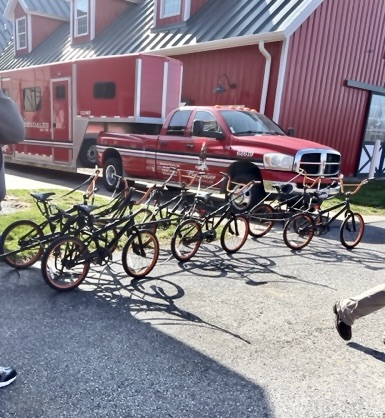 A set of six bicycles, paired two-by-two and harnessed together. Red pickup truck and red building behind.
